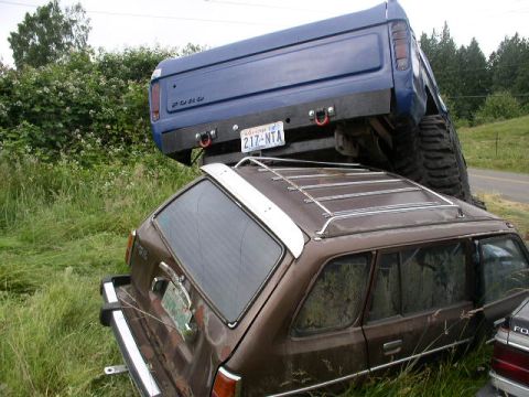 1979 Ford Bronco - A blue Ford pickup truck is precariously balanced on top of a brown station wagon in a grassy area.