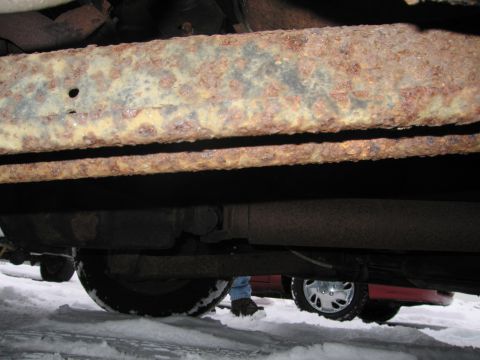 1991 Ford LTD Crown Victoria - The undercarriage of a vehicle shows significant rust on the frame rails, with snow on the ground and another vehicle in the background.