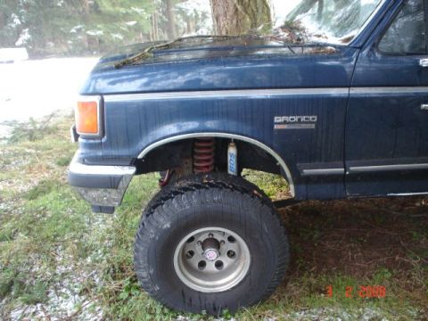 1990 Ford Bronco - A blue Ford Bronco is shown from the front quarter angle, large off-road tires and visible suspension modifications.
