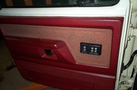 1996 Ford Bronco - Red vinyl door panel with a tan tweed cloth insert, molded armrest with integrated pull handle, and a black switch plate holding power wind…