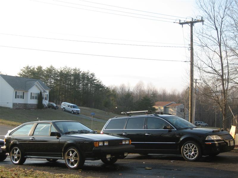 Black VW Passat wagon with roof rack parked beside a black coupe outdoors