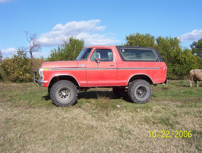 red Ford Bronco parked in a grassy field with trees and a cow in the background