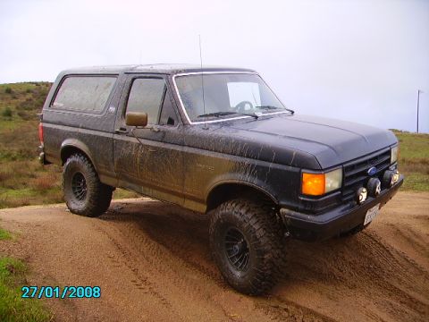 1990 Ford Bronco - 1990 Ford Bronco driving off-road on a dirt track, covered in mud.