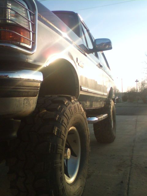 1995 Ford Bronco - A lifted Ford Bronco is viewed from the rear quarter panel with large off-road tires and chrome trim under a bright sun.