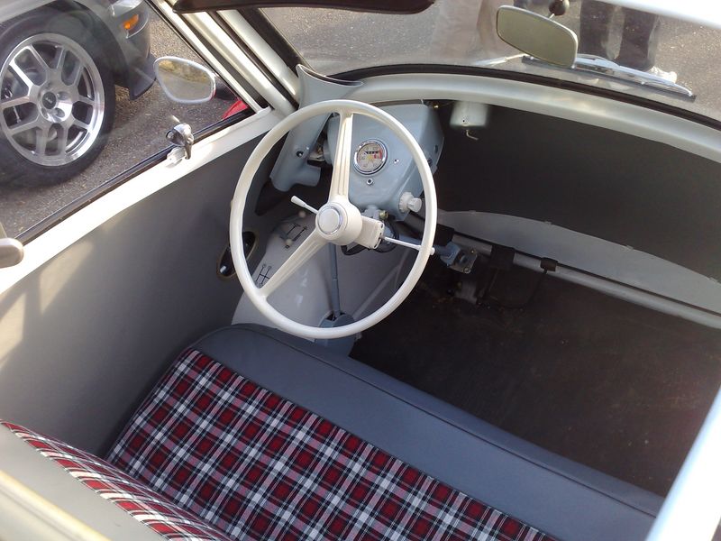 Interior of a vintage microcar with a white steering wheel and plaid seat. Another car's wheel is visible through the window