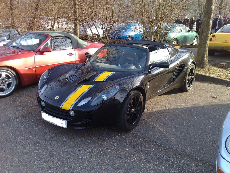 Black Lotus Elise convertible with yellow racing stripes, black wheels, parked outdoors