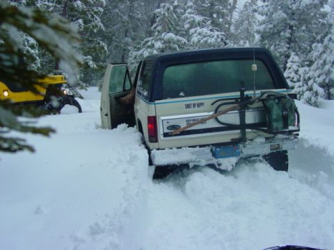 1986 Ford Bronco - A white 1986 Ford Bronco is stuck in deep snow, with a yellow vehicle partially visible behind it in a snowy forest.