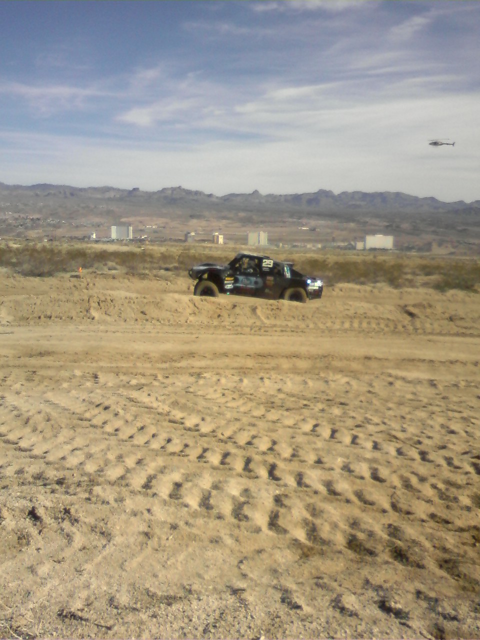 Off-road race truck with number 29 on door, driving on sandy terrain with helicopter overhead