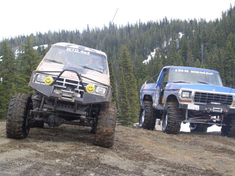 1979 Ford Bronco - Two modified trucks, including a 1979 Ford Bronco, are parked on a dirt path with a forest behind them.