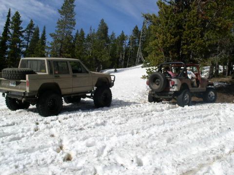 1979 Ford Bronco - Two modified off-road vehicles are parked in a snowy landscape with pine trees and a clear blue sky.