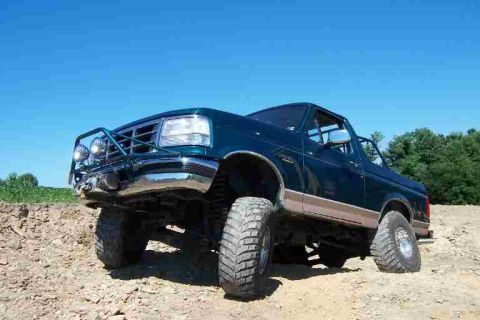 1996 Ford Bronco - Left front three-quarter view of a lifted teal-green Ford Bronco with the top and doors removed, showing a tubular front bumper with drivin…