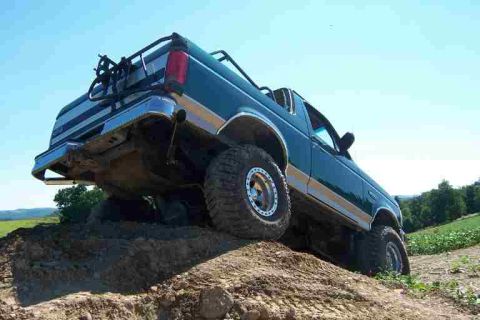 1996 Ford Bronco - Low rear three-quarter shot of a teal-green two-tone Ford Bronco with the hardtop removed, cresting a dirt mound so the rear tire lifts off…