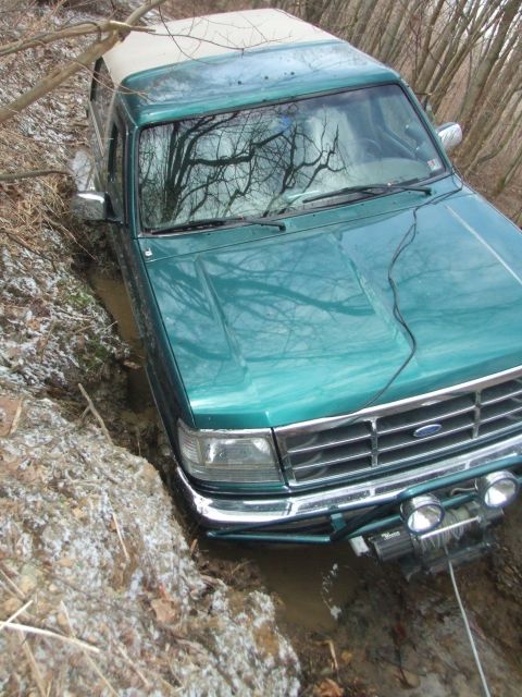 1996 Ford Bronco - High-angle view of a green Ford Bronco with a white hardtop bogged down in a muddy, water-filled rut in bare winter woods with patches of s…