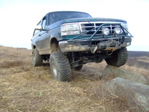 1996 Ford Bronco - Mud-splattered green Ford Bronco with the hardtop removed, straddling rocks in a field of dry winter grass with the right front tire off th…