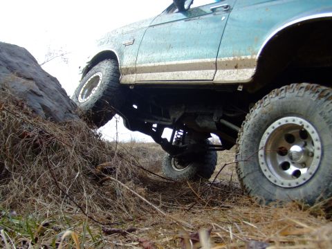 1996 Ford Bronco - Ground-level shot under the passenger side of a mud-caked green Ford Bronco perched on a rock, showing the lifted coil-spring front suspens…