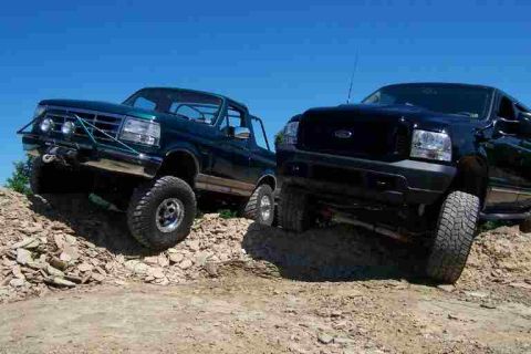 1996 Ford Bronco - A dark green lifted 1990s Ford Bronco with its top and doors removed, tube front bumper and black steel wheels on mud tires, parked on a sh…