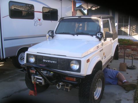 1985 Ford Bronco - White Suzuki Samurai with KC lights and winch, front view, as a person works underneath.