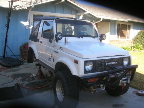 1985 Ford Bronco - White Suzuki Samurai on jack stands with aftermarket bumper and winch visible.