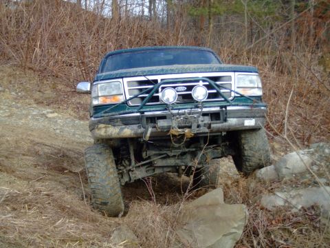 1996 Ford Bronco - Head-on view of a muddy green Ford Bronco with the top removed, articulating on a rutted dirt trail so the front axle is cross-flexed. A gr…