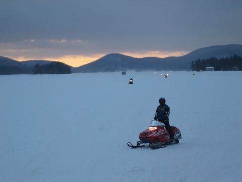 1995 Ford Bronco - A snowmobiler rides across a frozen lake at dusk, with other riders and distant mountains visible.