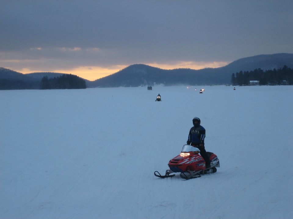 Person on red snowmobile on ice, mountains and sunset in background, other snowmobiles visible