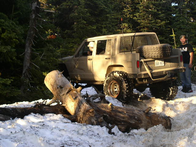 Tan Jeep Cherokee XJ with large tires driving over a fallen tree in a snowy forest
