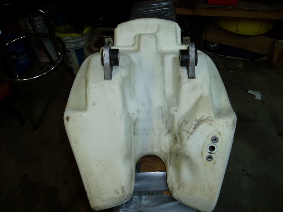 Plastic fuel tank with mounting brackets. It has a cutout in the middle and some mounting holes on the side