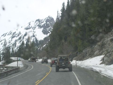 1979 Ford Bronco - A convoy of SUVs navigates a mountain pass with snow on the slopes and trees lining the road.
