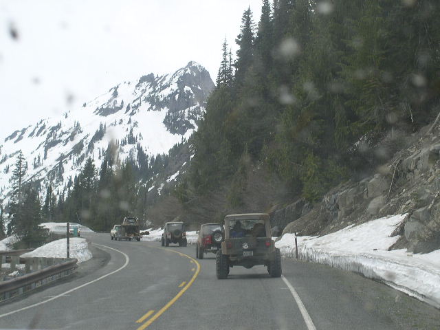 Several SUVs drive on a winding road with snow-covered mountains and trees