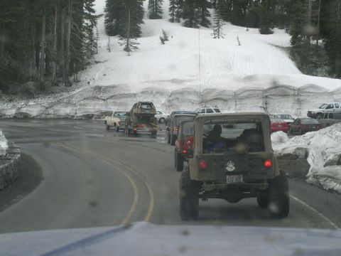 1979 Ford Bronco - Several off-road vehicles drive in a line on a paved road with snow banks and trees.