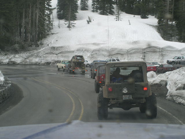 Line of off-road vehicles on a snowy mountain road, with trees and snow-covered hills
