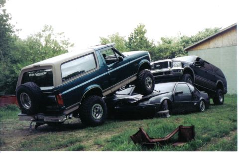 1996 Ford Bronco - Green Ford Bronco with a tan hardtop and a Tuff Country sticker in the rear glass climbing onto a crushed dark sedan in a grassy yard, with…