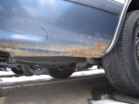 1991 Ford LTD Crown Victoria - The lower body panel of a blue car shows significant rust damage, with another vehicle visible in the background.