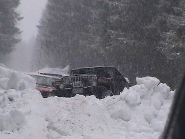 Dark Jeep Wrangler parked in deep snow amid falling snow and evergreen trees