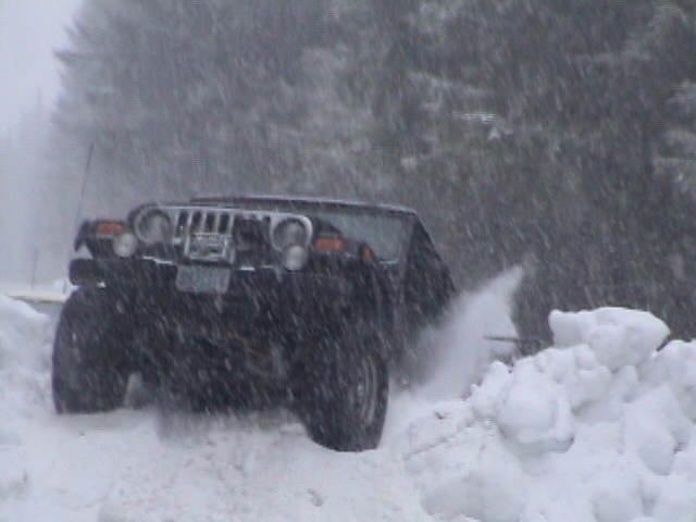 Dark Jeep Wrangler with light bar and big tires pushing through deep snow in a snowstorm