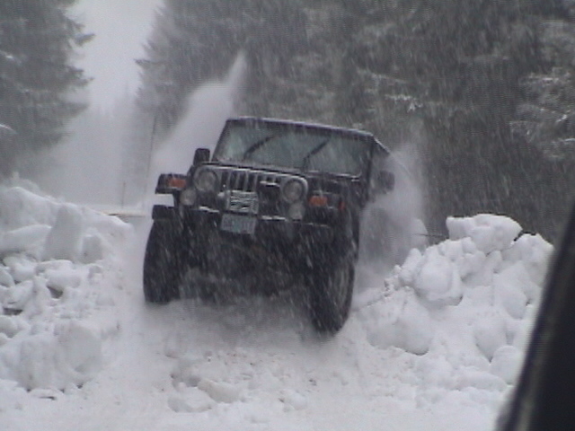 Dark Jeep Wrangler plowing head-on through deep snow on a forest road