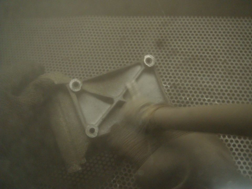 Metal bracket with four bolt holes and a pipe attached, seen in a dusty environment