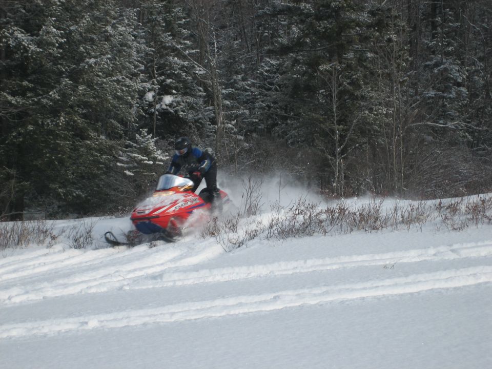 Person riding red snowmobile through snowy field with trees in background