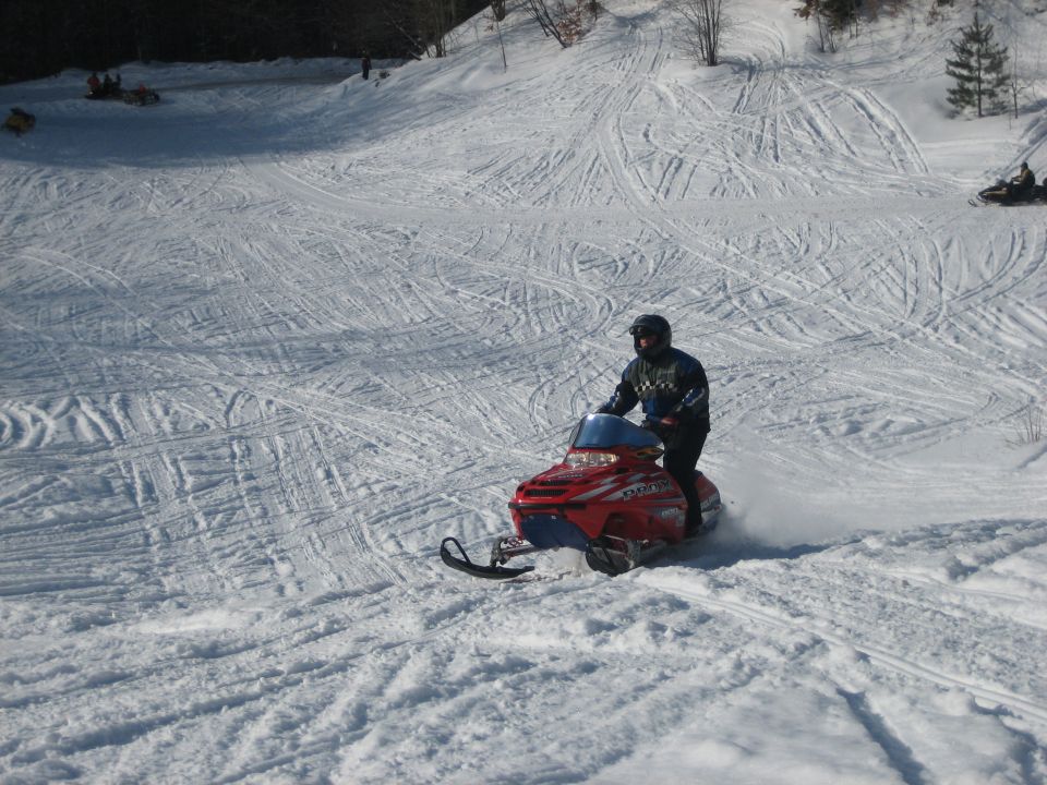 Red snowmobile with blue windshield and rider in helmet, angled left on snowy slope
