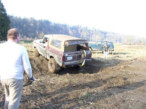 Mud-covered Ford Bronco backed into a muddy pit with a man and a green truck behind it