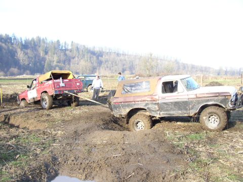 Brown Ford Bronco and a red truck stuck in deep mud ruts with people watching in a field