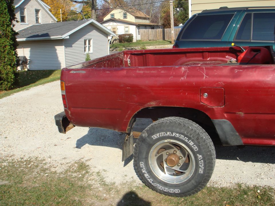 Dented red Toyota pickup bed on gravel with Goodyear Wrangler AT/S tire and chrome wheel