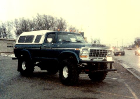 1979 Ford Bronco - This lifted pickup truck, possibly a 1979 Ford F-Series, features a white camper shell and off-road tires.