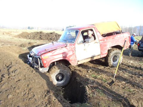 Red full-size 4x4 pickup with damaged front bumper stuck in a muddy off-road pit, driver inside