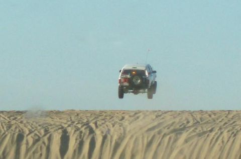 1991 Ford Bronco - An SUV is captured mid-air as it jumps over a sand dune against a clear blue sky.