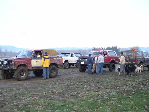 Row of lifted trucks and a red Ford Bronco parked in a muddy field with people and dogs