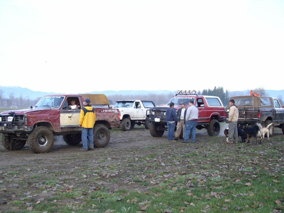 Row of lifted trucks and a red Ford Bronco parked in a muddy field with people and dogs