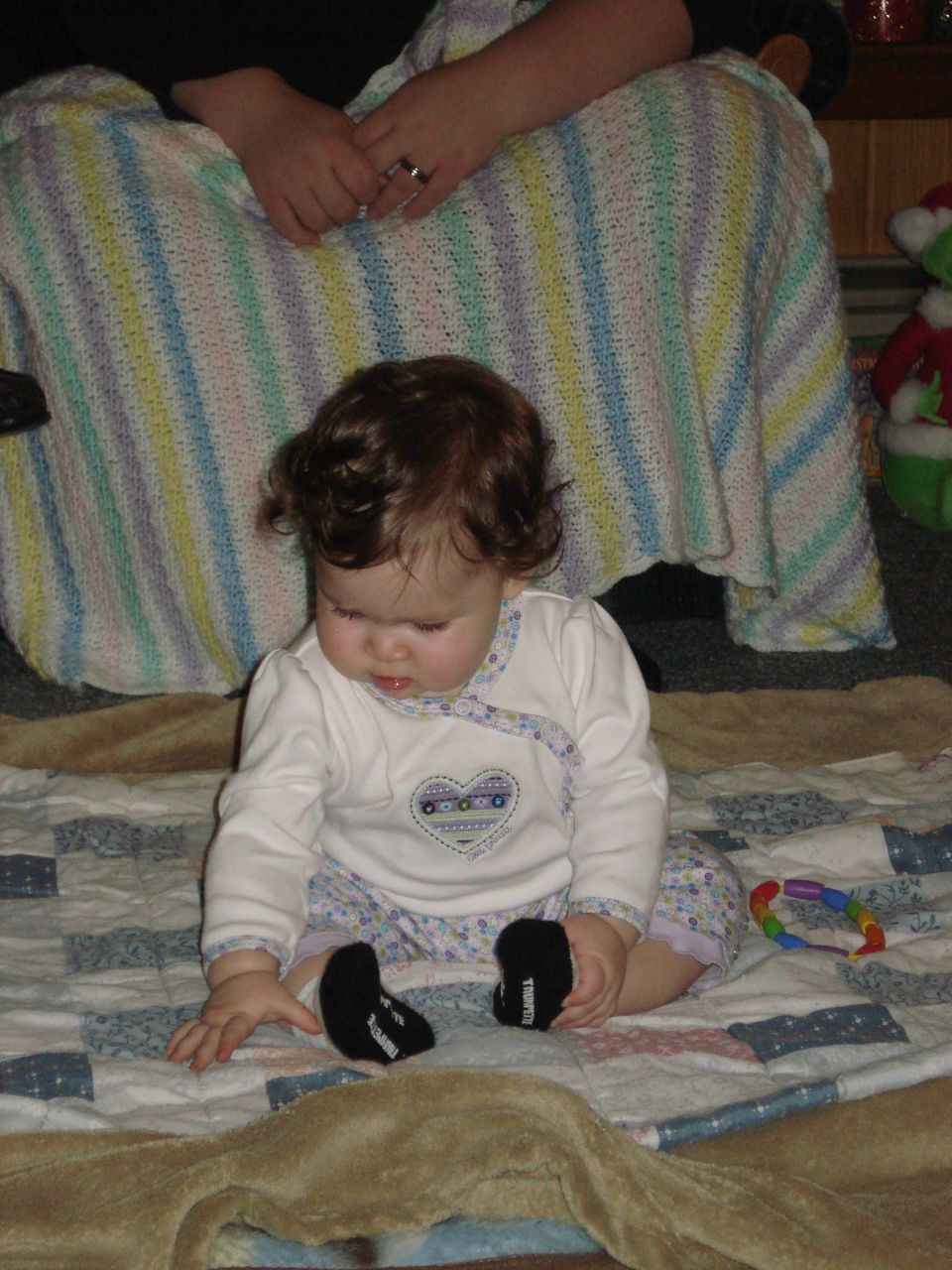 Baby sitting on a quilt with a toy nearby. Adult hands are visible behind the baby