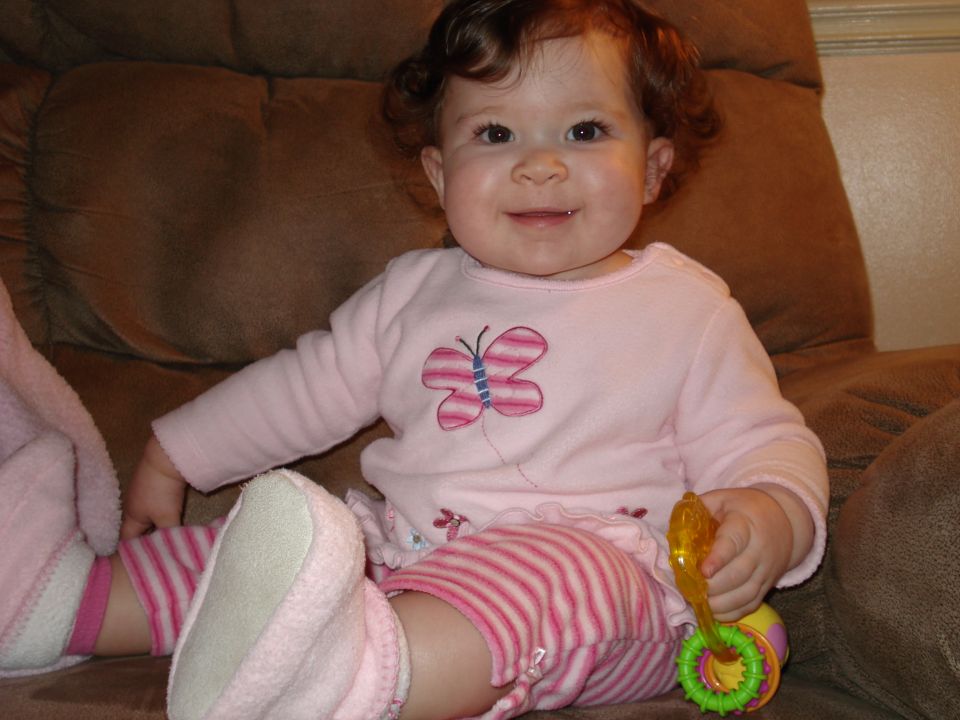 Baby girl with curly brown hair wearing a pink shirt with a butterfly and striped pants sits on a brown couch