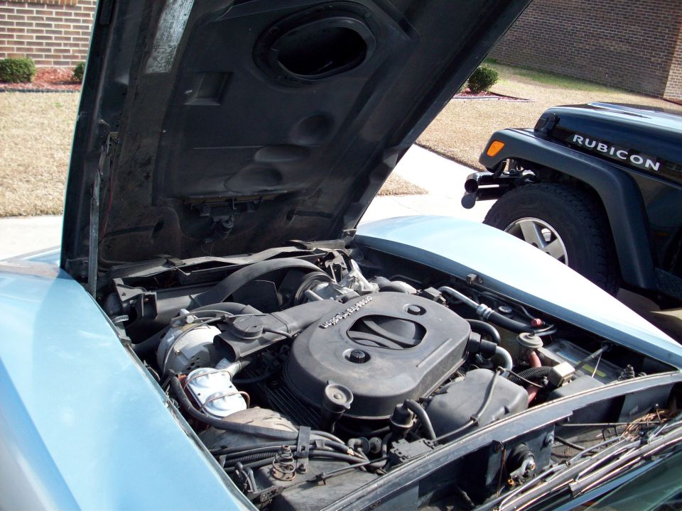 Engine bay with hood open, showing engine components. A Jeep Rubicon is visible in the background